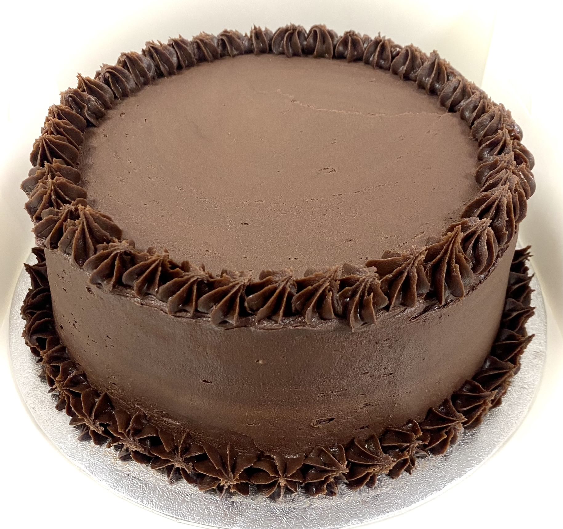 A round chocolate cake with smooth, rich chocolate frosting. The cake is decorated with piped chocolate icing in a rosette pattern along the top edge and base. The cake is presented on a silver cake board.